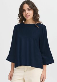 Top leggero blu navy con scollo a barca, maniche a 3/4 svasate e spacco laterale; texture liscia e dettagli minimi.