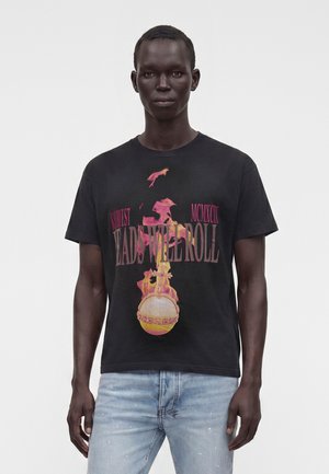 Mand iført en sort grafisk T-shirt med rød og gul design og tekst, der lyder "Heads Will Roll", kombineret med lyseblå jeans.