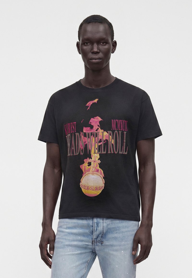 Άνδρας που φοράει μαύρο γραφικό T-shirt με κόκκινο και κίτρινο σχέδιο και κείμενο που αναγράφει "Heads Will Roll", συνδυασμένο με ανοιχτό μπλε τζιν.