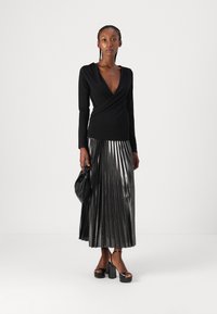 Top nero a maniche lunghe con scollo a V profondo, abbinato a una gonna pieghettata d'argento metallizzato. Modella tiene una borsa nera, indossando tacchi a piattaforma.