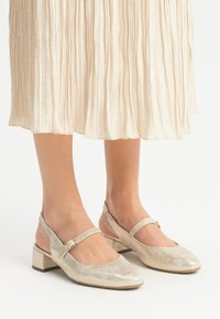 Scarpe slingback in pelle dorata con punta arrotondata, cinturino regolabile e tacco basso, abbinate a una gonna piegata di colore chiaro.