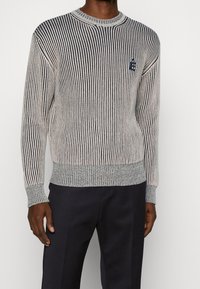 Strickpullover mit vertikalen navyblauen und cremefarbenen Streifen. Gerippter Rundhalsausschnitt und Bündchen, mit einem kleinen Logo auf der Brust. Textilstruktur.