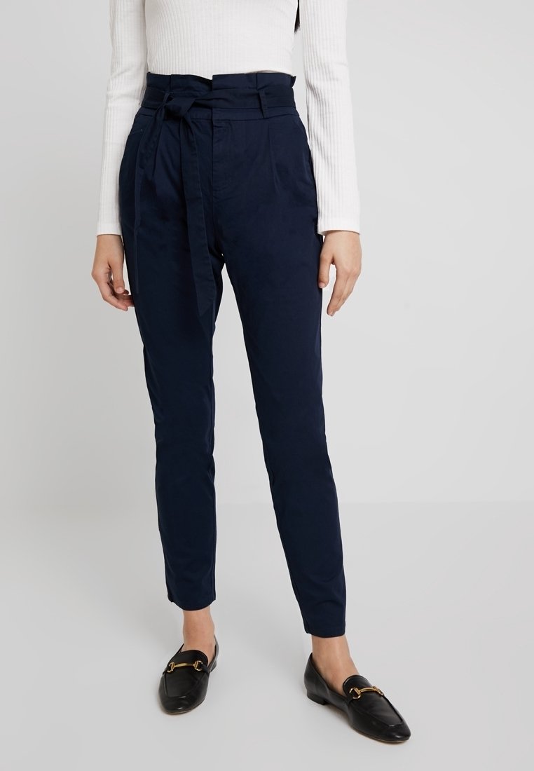 Pantalon bleu marine taille haute avec détail de ceinture nouée. Confectionné dans un tissu lisse, avec des jambes fuselées et des poches latérales.