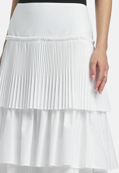 Kvindehånd hviler ved siden af en hvid lagdelt nederdel med tætsiddende og løse plisseringer mod en ensfarvet baggrund.