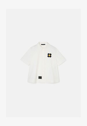 Camicia bianca a maniche corte con chiusura a bottoni e ricamo di un fiore nero. L'etichetta in basso include informazioni sul marchio.