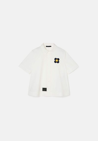 Witte shirt met korte mouwen, voorzien van een knoopsluiting en een zwarte bloemenborduursel. Label aan de onderkant bevat merkinformatie.