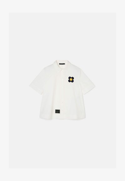 White short-sleeve shirt with a button closure and a black flower embroidery feature. Tag at the bottom includes brand information.