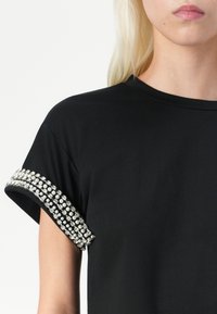 Black t-shirt with short sleeves featuring a decorative band of clear and pearl-like studs along the cuff. Smooth fabric, relaxed fit.