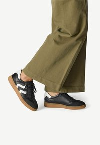 Baskets en cuir noir avec des accents en zigzag blanc, semelle en caoutchouc beige, et lacets noirs, associées à un pantalon large olive.
