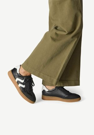 Sneaker in pelle nera con accenti zigzag bianchi, suola in gomma, e lacci neri, abbinati a pantaloni larghi verde oliva.