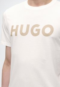 Κοντινό πλάνο με ανοιχτό μπεζ κείμενο «HUGO» σε λευκό κοντομάνικο μπλουζάκι που φοράει ένα άτομο.