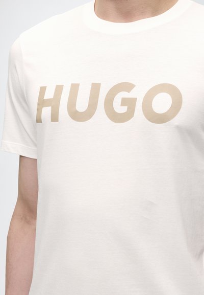 Крупен план на светлобежов текст "HUGO" върху бяла тениска с къс ръкав, носена от човек.