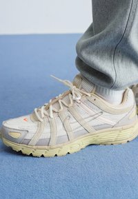 Beige joggesko med mesh og semsket skinn overdel, kremfargede snøringer og gul gummisåle. Merkbare oransje detaljer på tåen og siden.