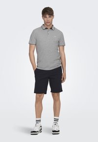 Graues Polohemd mit schwarzem Kragen und trim, kombiniert mit marineblauen Shorts. Modell trägt weiße Sneakers mit schwarzen Akzenten und gestreiften Socken.