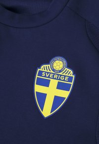 Σκούρο μπλε πουκάμισο με κίτρινη και μπλε σφραγίδα της Σουηδικής Ομοσπονδίας Ποδοσφαίρου, με σχέδιο ασπίδας, υφή στην λαιμόκοψη και απαλό ύφασμα.