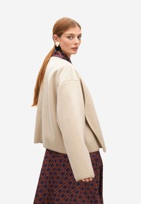 Mujer con cabello largo y rojo recogido, lleva una chaqueta beige de gran tamaño sobre un vestido estampado en marrón y azul marino, de perfil sobre un fondo blanco.