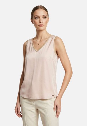 Donna che indossa una camicetta rosa chiaro senza maniche con rifiniture dorate e pantaloni beige, in piedi e che guarda leggermente di lato su uno sfondo bianco.