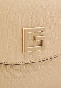 Beige håndtaske med et præget mønster, der har en firkantet logo i guldfarve og syninger langs kanten.