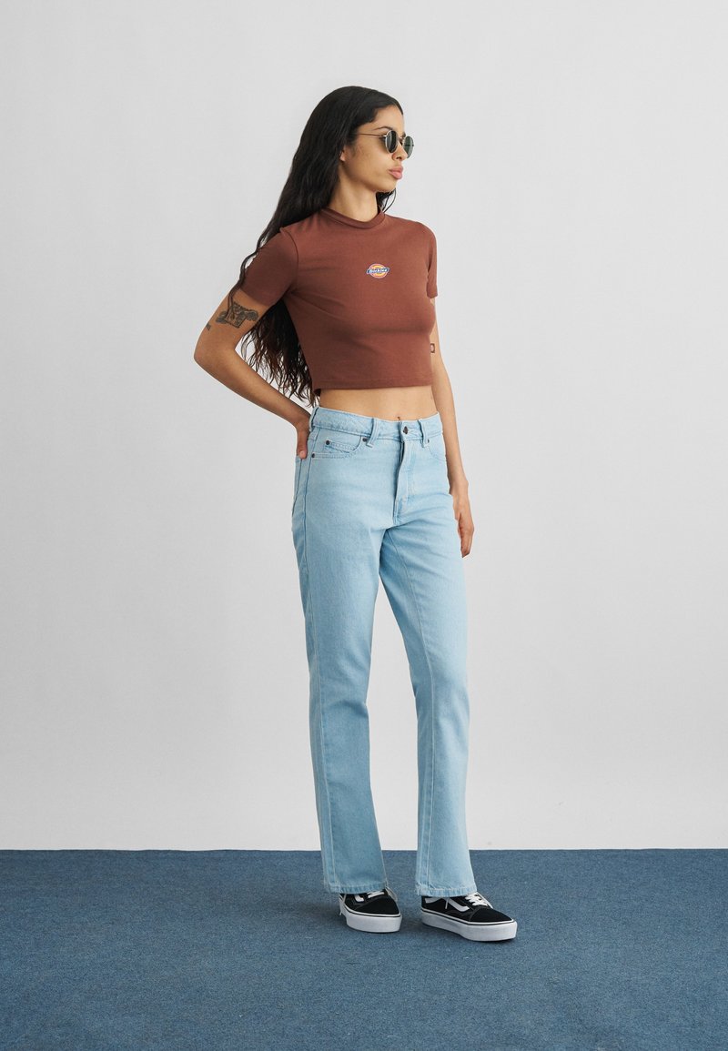 Junge Frau mit langen dunklen Haaren, die ein braunes kurzes T-Shirt, hellblaue Jeans, schwarze Sneakers und Sonnenbrille trägt, steht auf einem blauen Teppich.