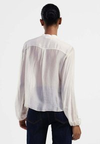 Blusa leggera e trasparente di colore bianco, con maniche lunghe arricciate e scollatura rotonda, caratterizzata da un dettaglio cucito sul retro e una vestibilità comoda.