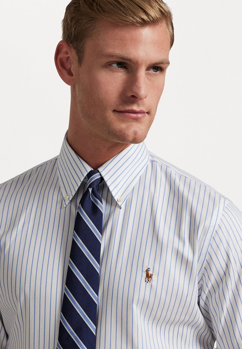 White dress shirt with light blue vertical stripes, button-down collar, and a small embroidered logo. Paired with a navy and white striped tie.