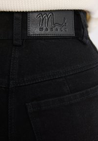 Czarne dżinsy z denimu z tylną kieszenią i czarną skórzaną metką z napisem "Makalu", noszone z kremowym swetrem z dzianiny.