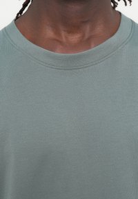 Ανοιχτό πράσινο, κοντομάνικο μπλουζάκι με στρογγυλή λαιμόκοψη. Κατασκευασμένο από μαλακό ύφασμα με λεία υφή, διαθέτει διακριτική, ομοιόμορφη ραφή κατά μήκος της λαιμόκοψης.