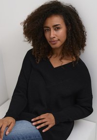 Schwarzer gerippter Strickpullover mit V-Ausschnitt und asymmetrischem Saum, kombiniert mit blauen Jeans. Das Modell hat lockiges Haar und legt eine Hand auf ihren Schoß.