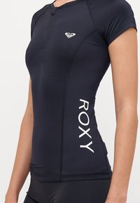 Navy kortermet rash guard laget av elastisk stoff, med halv glidelås og hvit "ROXY"-logo på siden. Glatt tekstur og tettsittende design.