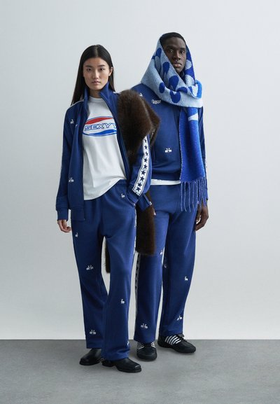 Blå träningsdräkter med vita stjärnmönster, en grafisk vit t-shirt och en texturerad blå scarf. Modellerna bär olika svarta skor.