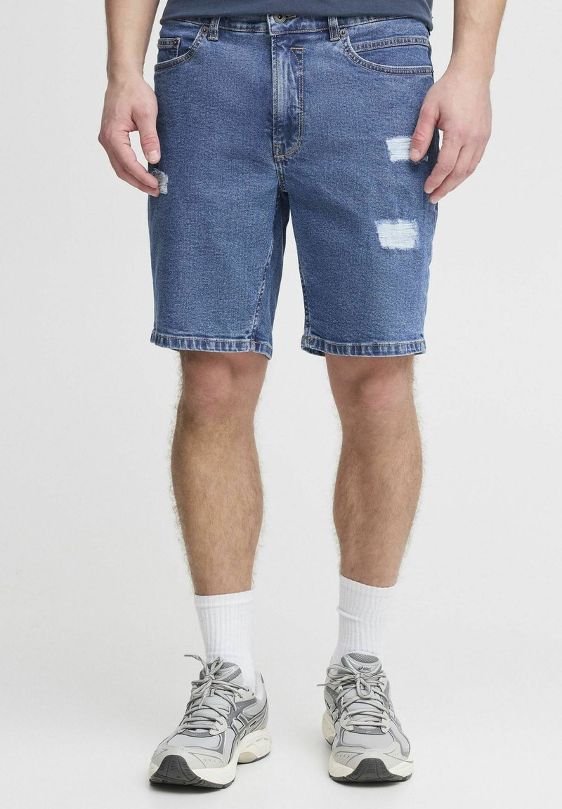 Shorts de mezclilla en azul medio con bordes deshilachados, que presentan dos parches desgastados. Con un corte recto y cinco bolsillos.