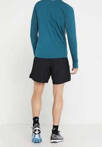 Teal lange mouwen sportshirt gecombineerd met zwarte shorts. De outfit bevat gestreepte schoenen met blauwe accenten.