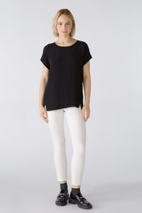 Schwarzes, kurzärmliges Oberteil aus glattem Stoff, kombiniert mit weißen, taillierten Hosen. Das Outfit umfasst glänzende schwarze Schuhe und dezente Details.