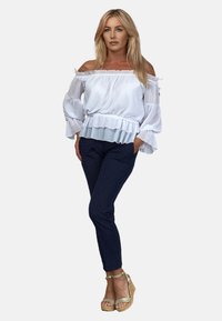 Biała bluzka bez ramiączek z falbaną, zestawiona z ciemnoniebieskimi dopasowanymi spodniami i beżowymi sandałami na koturnie. Gładki materiał, elegancki fason.