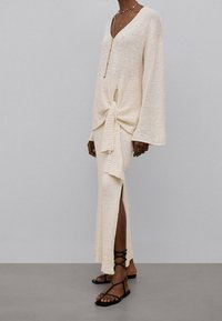 Robe en tricot crème avec des manchons en forme de cloche et un décolleté en V profond ; détail de nœud noué à la taille ; fente sur le côté ; associée à des sandales noires à lanières.