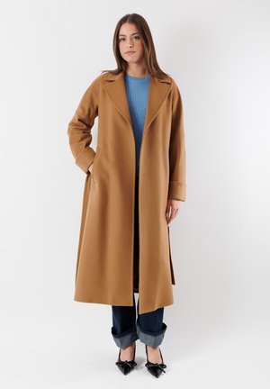 Cappotto di lana marrone con colletto a taglio e maniche lunghe, indossato sopra un maglione blu a coste, abbinato a jeans di denim scuro con risvolto e tacchi neri.