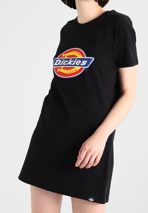 Osoba ubrana w czarną sukienkę z krótkim rękawem z dużym, kolorowym logo Dickies na klatce piersiowej na jednolitym jasnym tle.