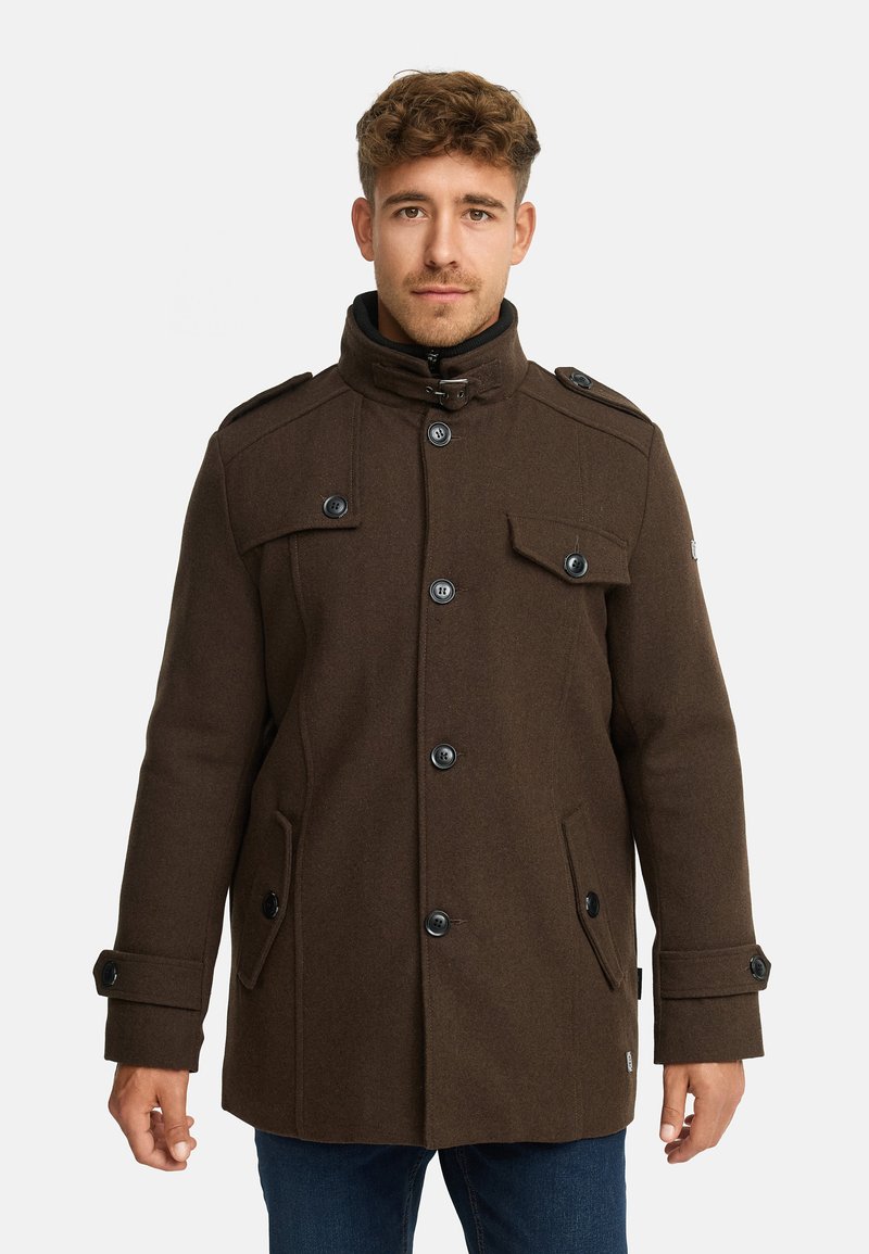 Brun frakke i uldblanding med høj krave, dobbeltradet design, store knapper og klaplommer. Har skulderstropper og en struktureret form.