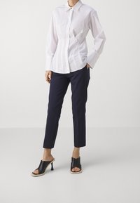 Chemise blanche à manches longues avec un design structuré, associée à un pantalon fuselé bleu marine foncé et des sandales à talons noirs ouvertes à enfiler.