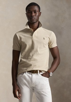 Νέος άνδρας φοράει μπεζ πόλο με όρθιο γιακά, λευκό παντελόνι, καφέ ζώνη και καφέ ρολόι σε ουδέτερο φόντο.