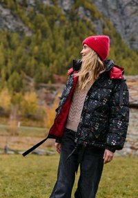 Kurtka puchowa z czarnym kwiatowym wzorem i czerwoną podszewką, z wysokim kołnierzem i regulowanym pasem. Połączona z beżowym swetrem dzianinowym i ciemnymi spodniami.