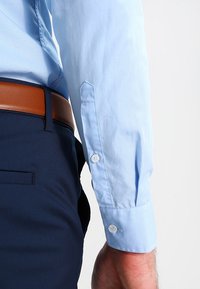 Ljust blå knapparskjorta med en slät textur, med två vita knappar på ärmsluten, kombinerad med mörkblå byxor och ett brunt bälte.