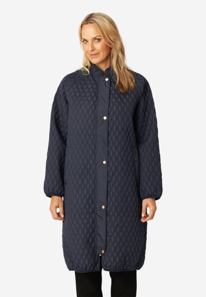 Donna che indossa un lungo cappotto trapuntato blu navy con bottoni a pressione, in piedi davanti a uno sfondo chiaro uniforme.