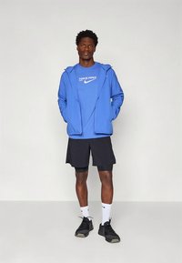 Blauw sportjack over een blauw Nike Pro-shirt, zwarte shorts en zwarte schoenen, met witte sokken. Model staat met de handen in de zakken.