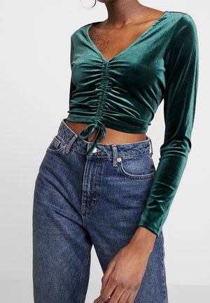 Persoon die een donker teal fluwelen langärmelig gerimpeld cropped top met een strik en een hoge taille blauwe spijkerbroek draagt tegen een effen achtergrond.