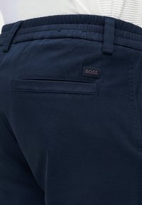 Σκούρο μπλε παντελόνι με πίσω τσέπη με εσοχή και ετικέτα με το λογότυπο «BOSS», φορεμένο με ένα λευκό υφασμάτινο πουκάμισο που έχει μπαρτωθεί μέσα.