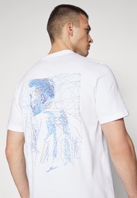 Hvit bomulls t-skjorte med et blått og brunt geometrisk linjekunstdesign på baksiden, som viser en stilisert figur og signert detalj.