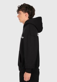 Un jeune homme aux cheveux bouclés portant un sweat à capuche noir avec du texte blanc, se tenant de profil contre un fond clair.