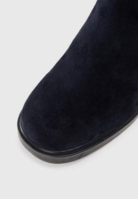 Navy suede nilkkurit, joissa on sileä pinta, pyöreä kärki ja musta kumipohja. Saumaton muotoilu ilman näkyviä metalliosia tai koristeita.