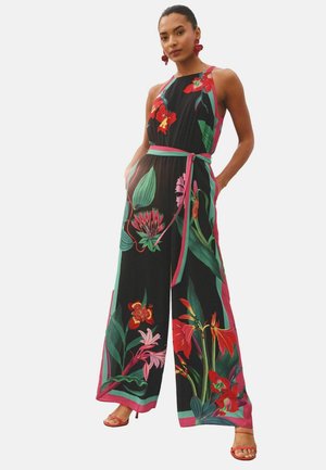 Woman wearing a sleeveless black jumpsuit with vibrant red and green floral prints, a tied waist belt, and red high-heeled sandals.